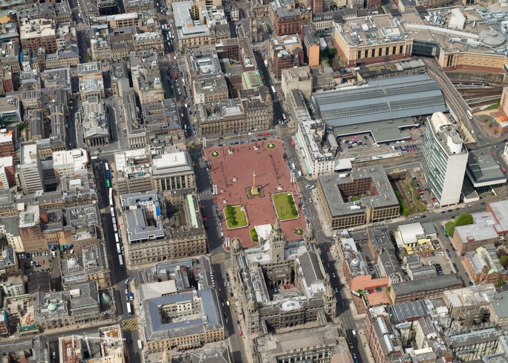 A bird's eye view of a city, focused on a large square with a red floor and a tall statue in the centre