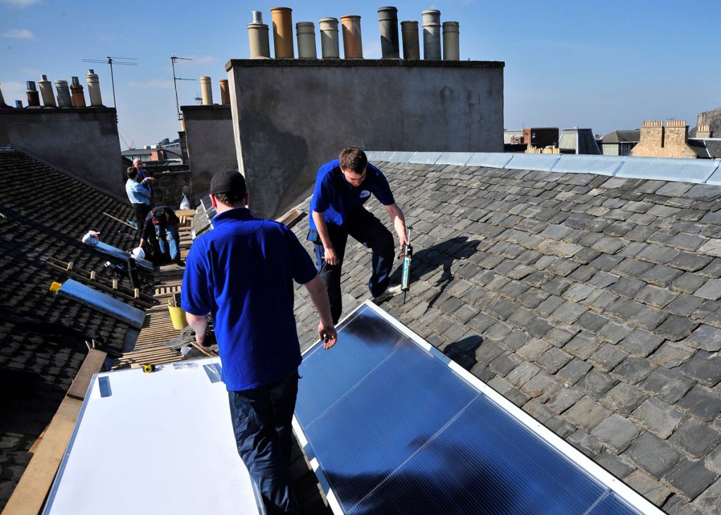 Two men stand on a rooftop securing solar panels to tiles. Chimney pots can be seen in the distance.