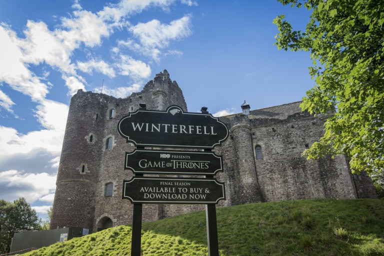 A black sign reading "Winterfell" in front of the imposing walls of Doune Castle