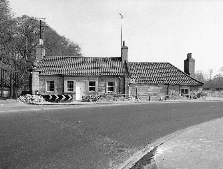 Black and white picture of a small cottage on the corner of a road approaching a bridge 