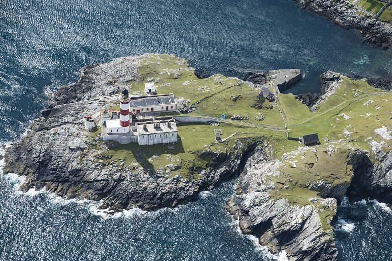 An aerial photo of a lighthouse on a small piece of land surrounded by the sea. The land near the lighthouse is visibly eroding