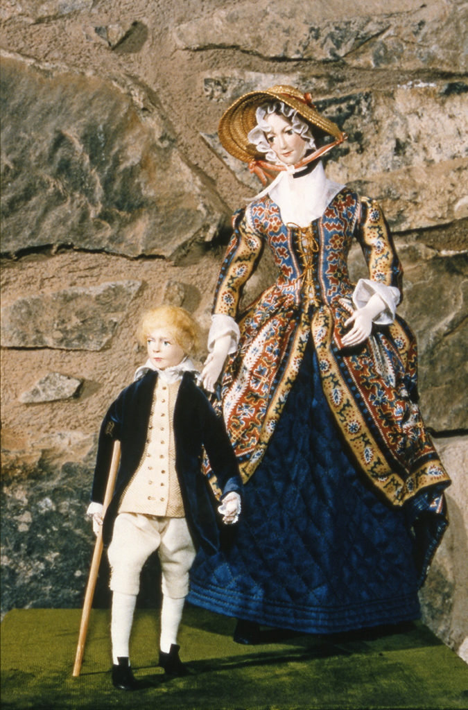 Doll of a woman dressed in a fine dress standing beside a child wearing a frock coat and carrying a stick.
