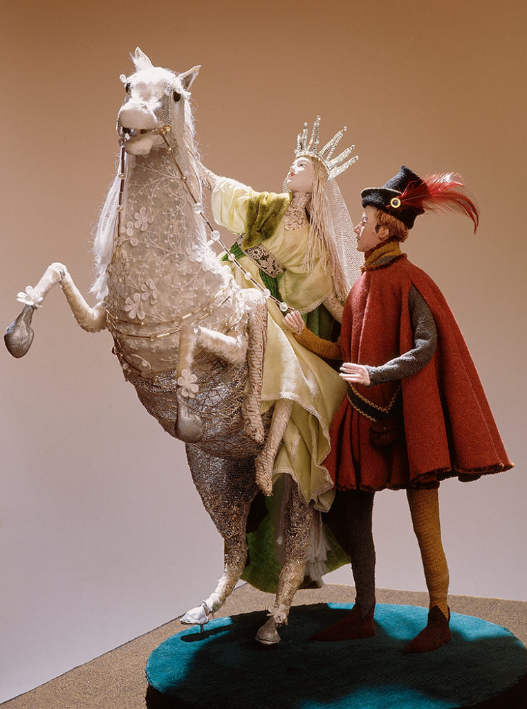 A woman wearing a crown and a fine dress sits on rearing horse. Beside her stands a man in a red cloak wearing a hat with a red feather in it.