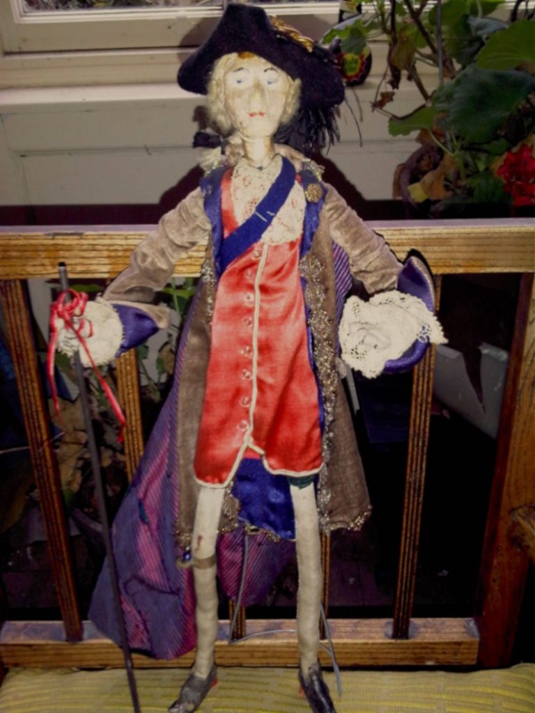 A doll wearing a red waistcoat, long cloak and a tricorne hat.