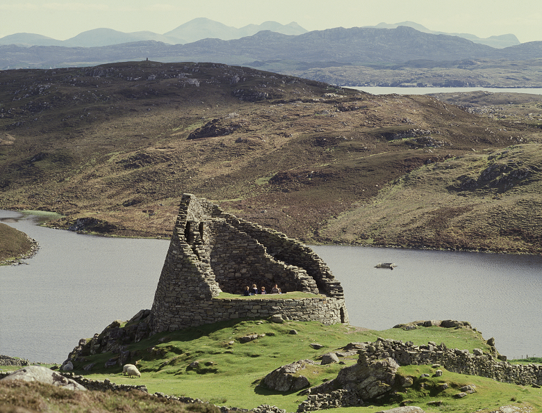 View of a broch on a rocky outcrop in front a small loch with the sea beyond