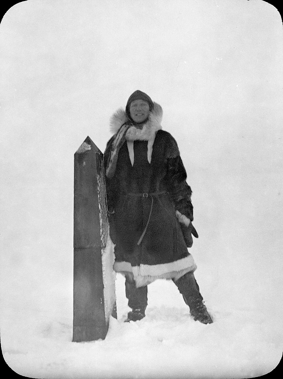 Blakc and white photo of Isobel Hutchison in Artic exploration gear posing at a small monument marking the border between the USA and Canada