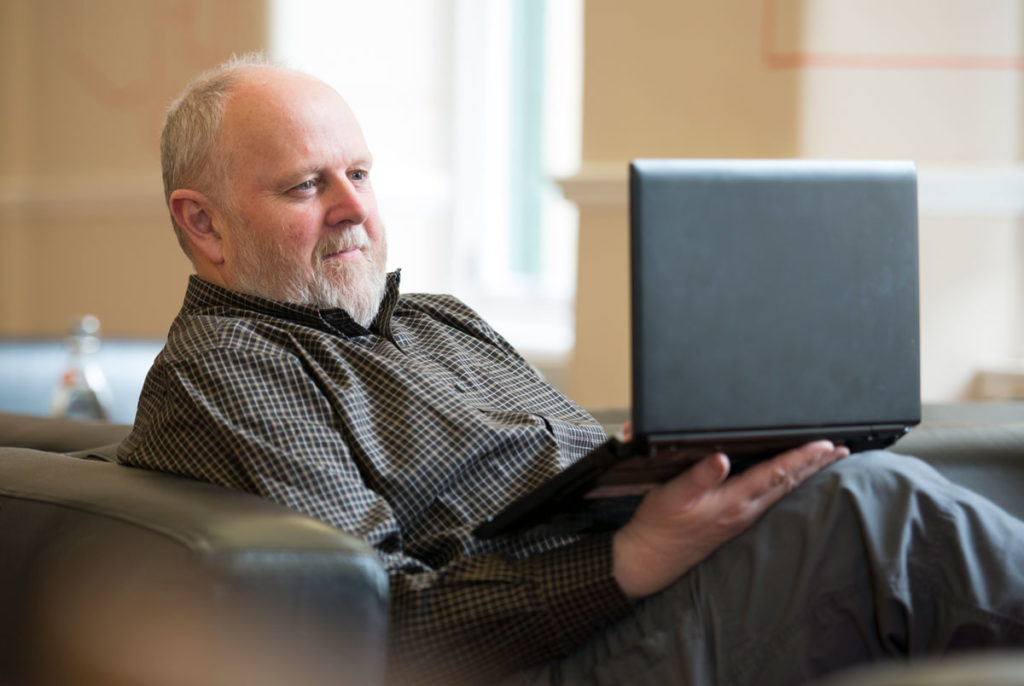 A man with a white beard sits on a sofa and looks at a laptop, smiling