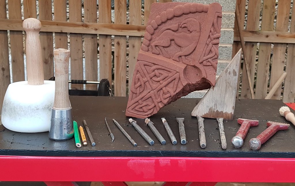 Stonemason's tools on a bench surrounding a carved replica of a Pictish stone