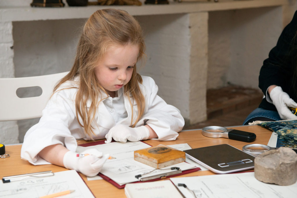 A girl in a white coat and wearing white gloves looks at an object in front of her as she holds a pencil