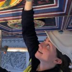 A woman wearing a hard hat works on a painted ceiling.