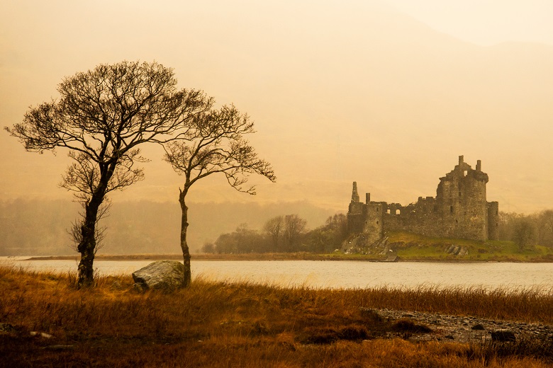 An atmospheric photo of two trees silhouetted against Loch Awe and Kilchurn Castle