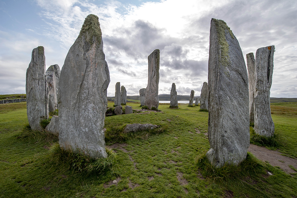 view of Calanais Standing Stones, a circle of tall grey stones of different sizes on green grass with cloudy sky above