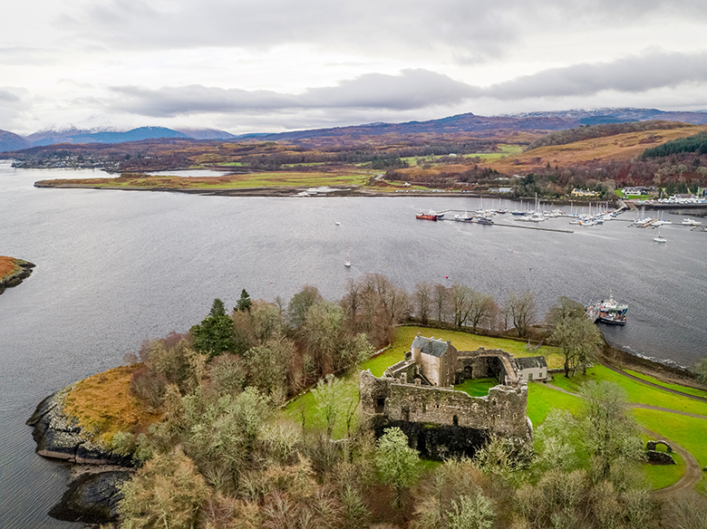 Aerial view of the ruins of Dunstaffnage Castle. It sits close to the shore surrounded by trees.