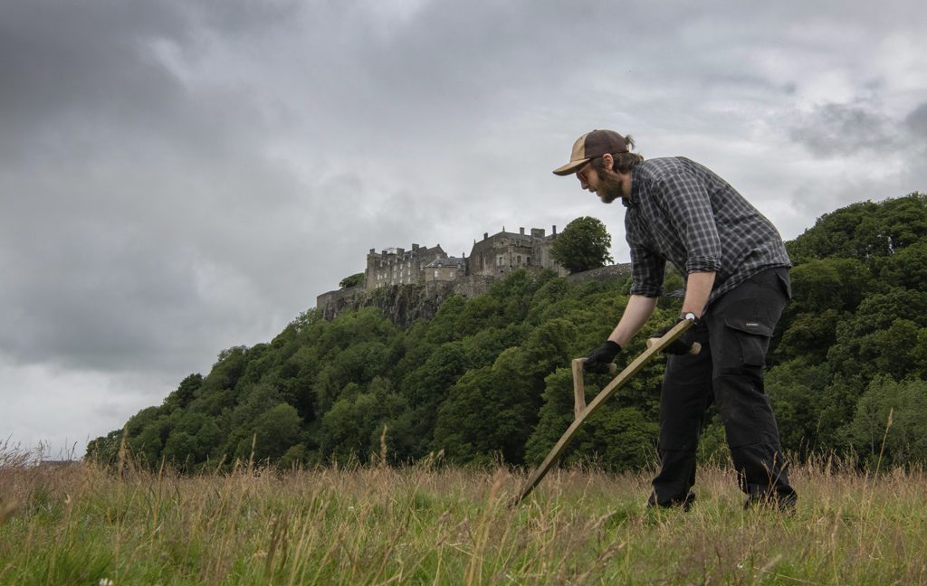 man in hat and gloves carries out scything, a traditional method of grass cutting using a curved blade which dates back to ancient times