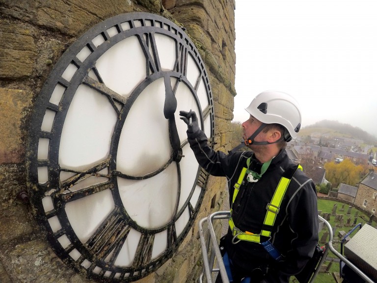 close up of a steeple clock specialist wearing hard hat and safety gear working on one of the faces of a church clock on the side of a tower