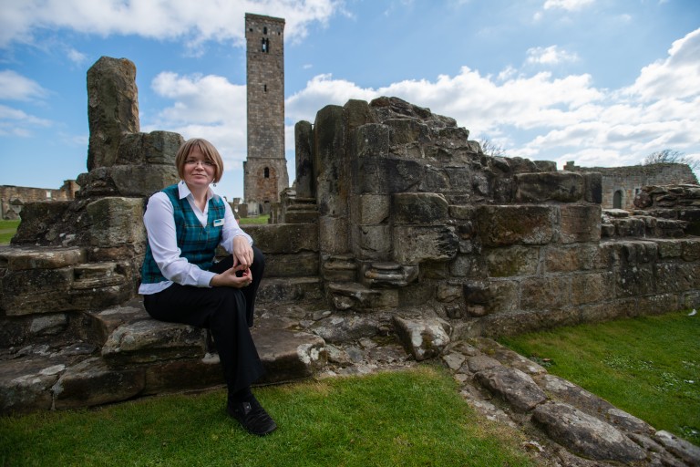 woman in white shirt, black trousers and tartan waistcoat sits on a wall with a ruined building and a tall tower visible behind her