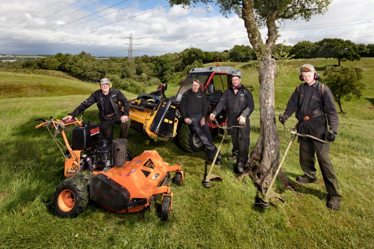 four men standing by a tree holding various gardening equipment face the camera