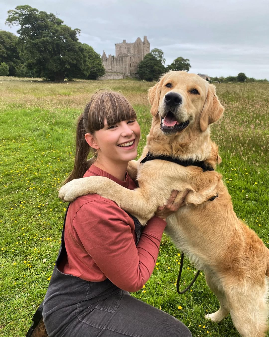A smiling golden retriever stands on his hind legs with his paws around his owner's shoulders, looking at the camera. There's a ruined castle in the background.