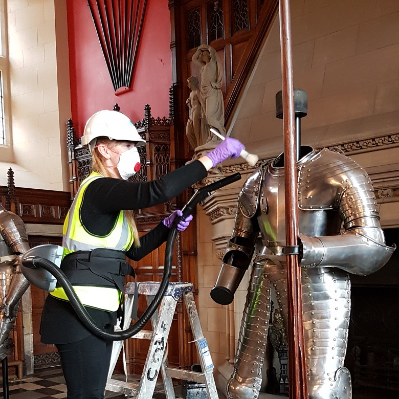 A masked and helmeted conservator cleans a suit of armour