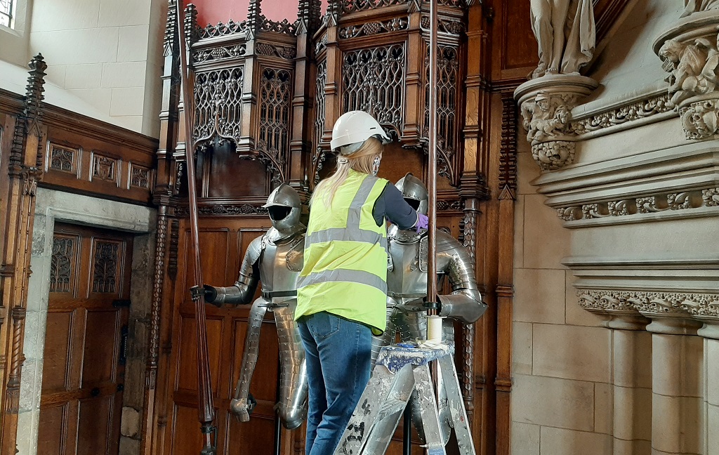 A conservator in a mask, helmet and high-vis jacket stands on a step ladder in order to clean a suit of armour