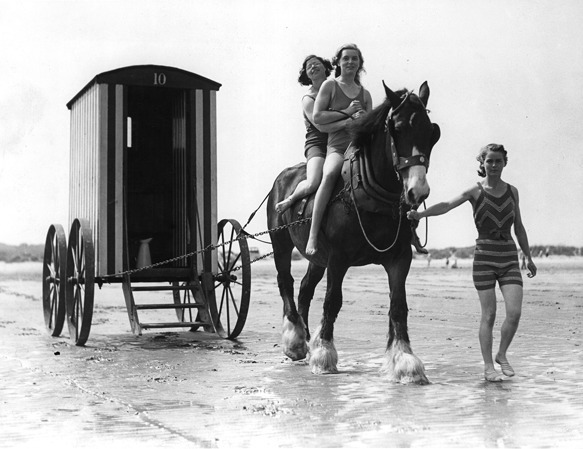 Horse carrying people in swimming costumes hauls a bathing-hut along the sand to the sea