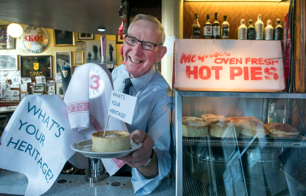 A smiling man stands behind the bar and offers a pie