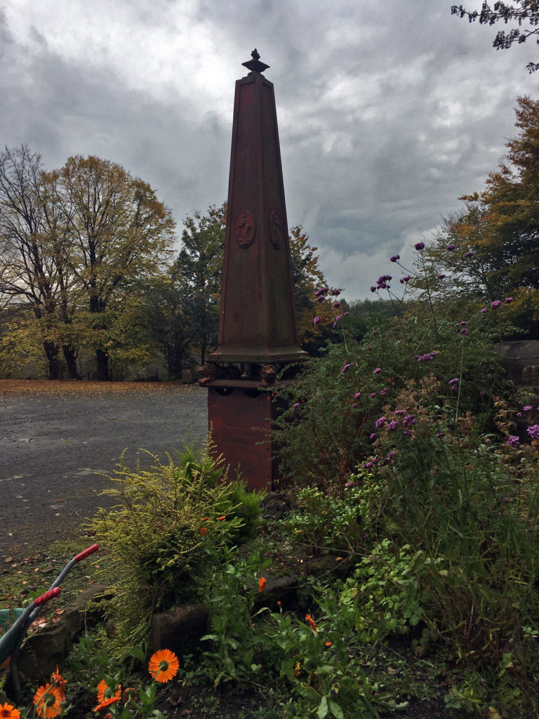 A tall cast iron obelisk by some trees and bushes