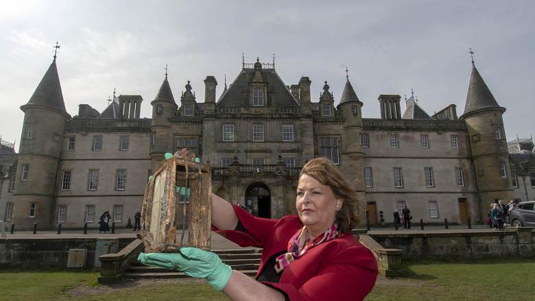 A lady in front of a large stately home holding a metal lantern while wearing gloves