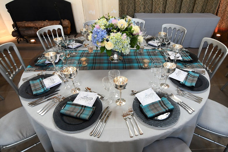 A circular table laid out for a gala dinner featuring a centrepiece of colourful flowers, tartan napkins and tablecloths, silver wine glasses and candles