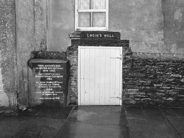 A black and white photo of a sign reading "Login's Well" above a white wooden doorway on the side of a house. To the left of the door is a stone plaque listing vessels which have used the well.