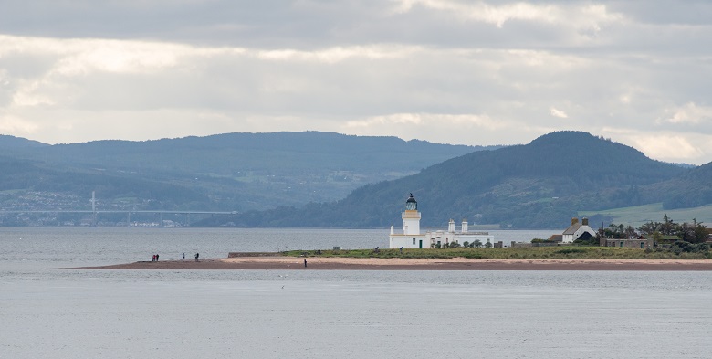 A white lighthouse on a promontory with mountains and the faint outline of a bridge in the background