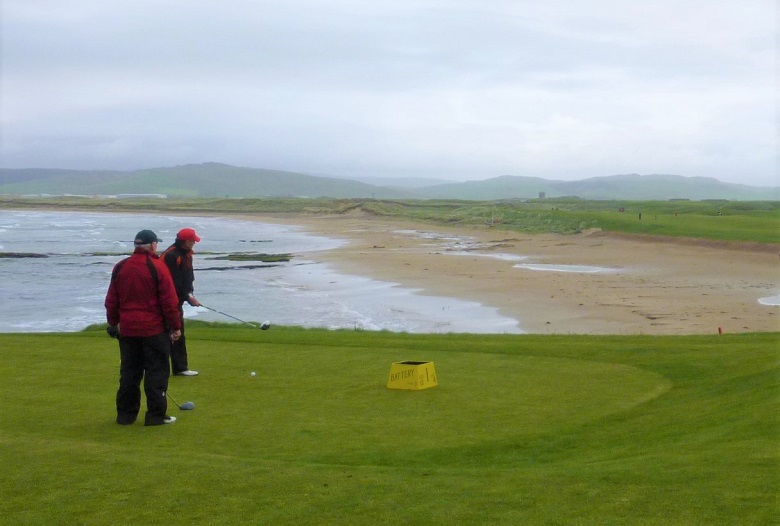 Two golfers teeing off on a windy day with the sea in the background