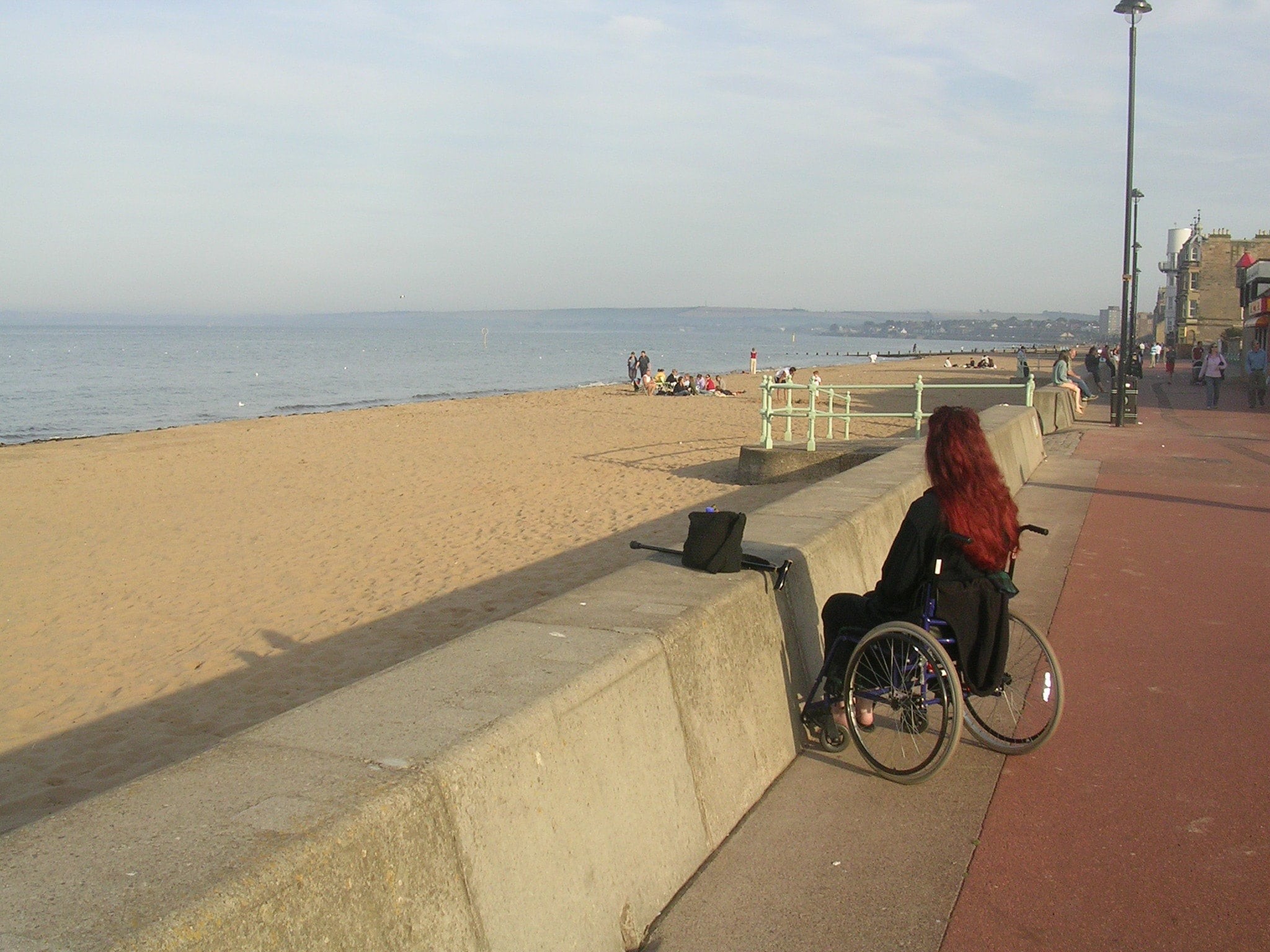 A person in a wheelchair looks out to sea from an urban promenade. There is a beach between the promenade and the water but no access path can be seen