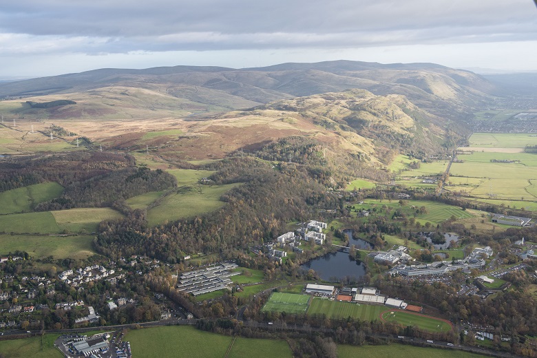 An aerial view of modern university buildings set into green fields and countryside with mountains in the background 