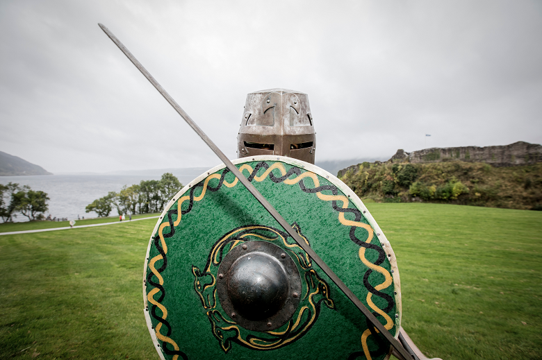 A living history performer wearing a metal helmet and holding a long sword and a round, green shield