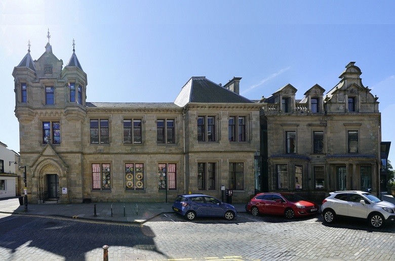 Cars parked in front of an ornate stone library building in a Scottish town centre. A small tower with turrets is above the main entrance to the building. 