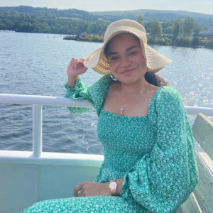 A woman from a mixed ethnic background sits on a boat in a flowery dress and sun hat, smiling at the camera