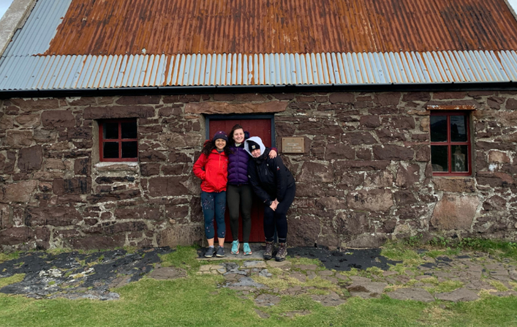 three young women in outdoor wear stand outside a bothy