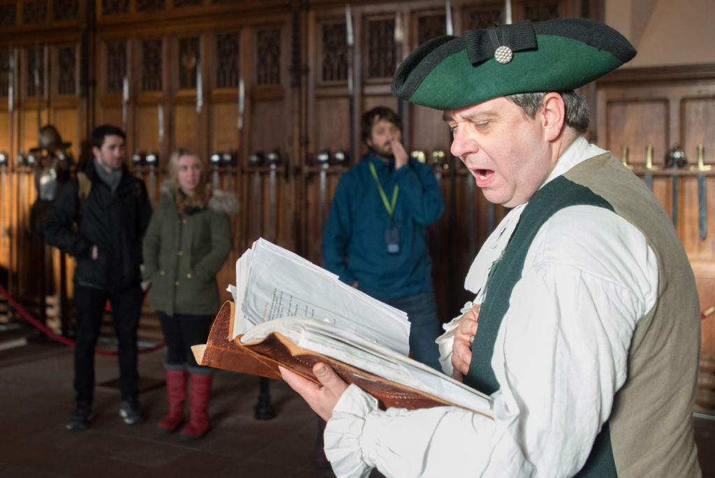 A man dressed as Rabbie Burns reads from a book, speaking loudly to an audience