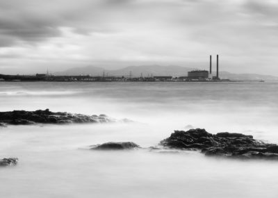A black and white photograph of a sea landscape with a power station in the background.