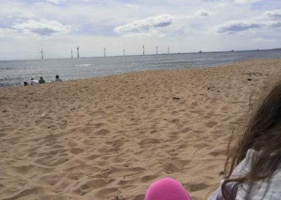 A young girl sits on the beach, which overlooks wind turbines