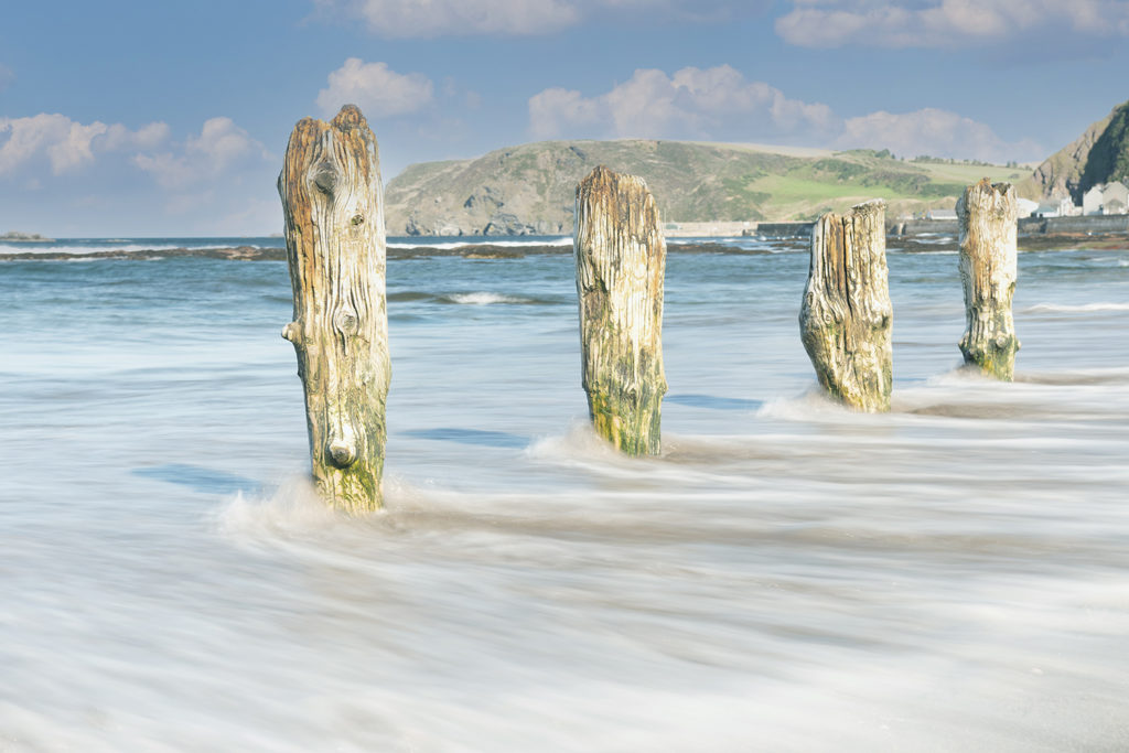 Old jetty posts stand rotten on a beach. The sea flows between them.