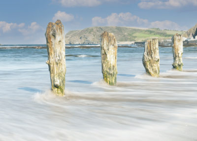 Old jetty posts stand rotten on a beach. The sea flows between them.