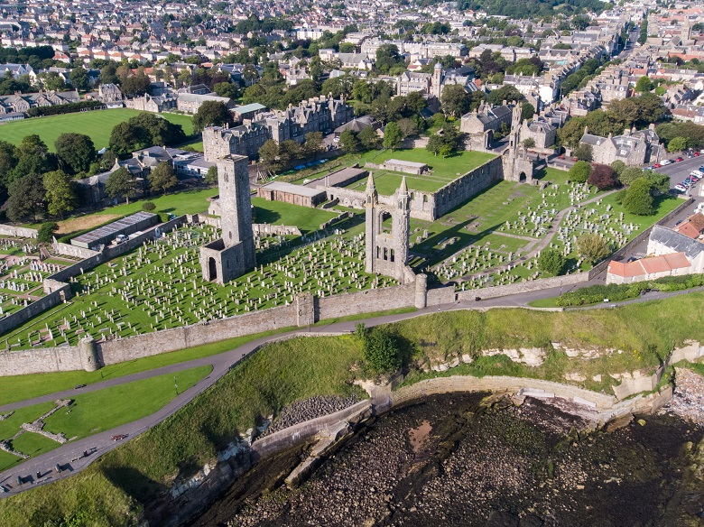 An aerial view of the ruins of a large cathedral surrounded by a graveyard and a stone perimeter wall 