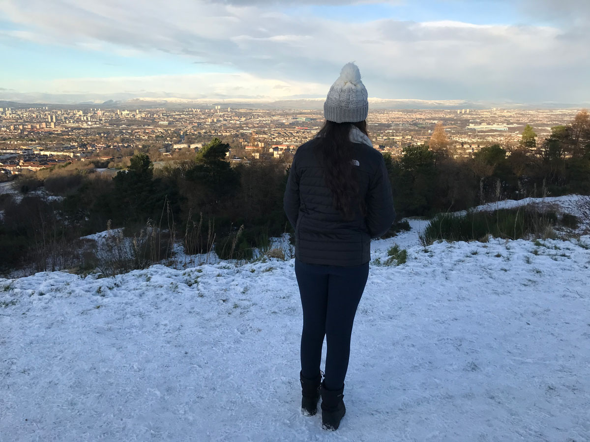 A teenager wearing a bobble hat stands on a snowy hill and looks out across the city