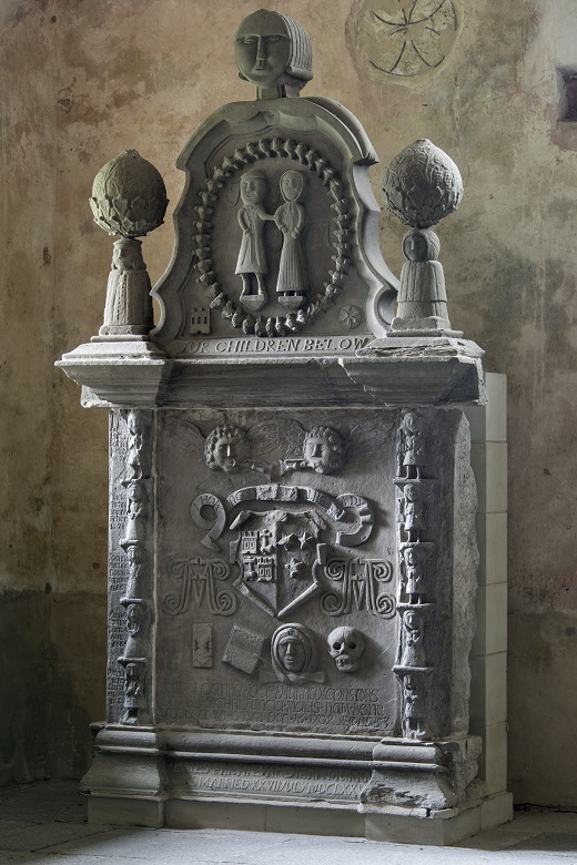 An intricately carved gravestone featuring a central carving of a husband and wife standing arm in arm. Below there are several other carvings depicting the couple's children and interests