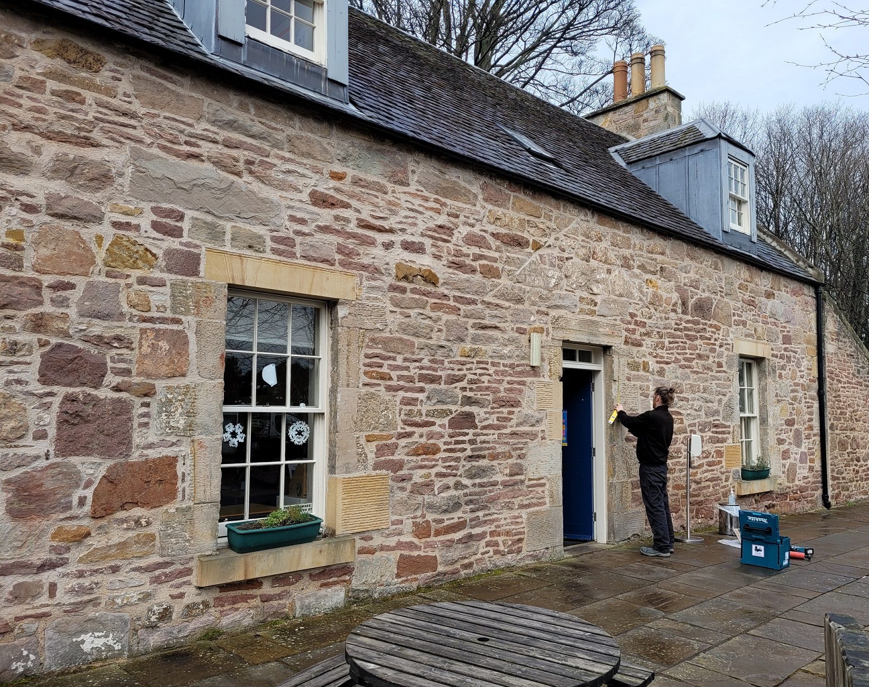 Photo outside of Liberton bank House with a person drilling a hole in the wall and another person in high-vis jacket standing by.