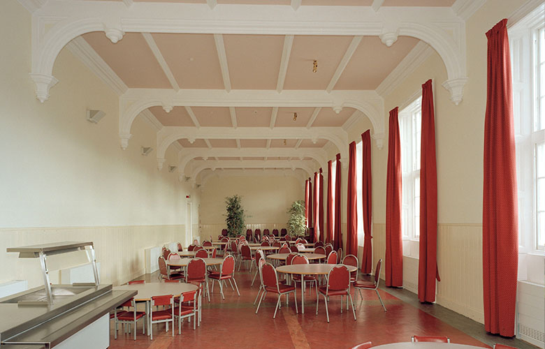 A large but utilitarian looking dining room. There are fllor to ceiling windows on the right hand side of the room. There are 7 or 8 large circular tables surrounded but 5 chairs each. 