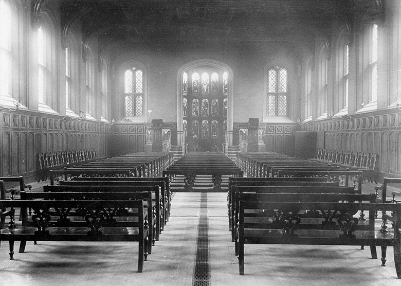 a black and white photo of a large chapel with two columns of pews, carved wooden panelling on the lower half of the walls and windows along each wall. There is an ornate window behind the altar.