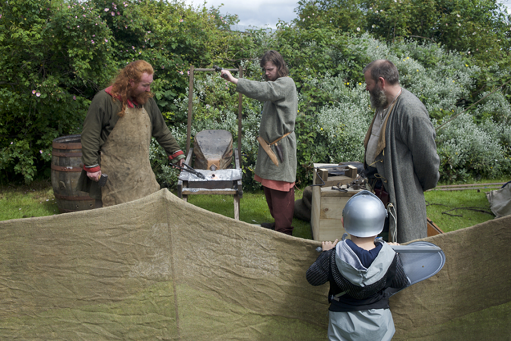 Photo of 3 people dressed in reenactment clothing working on metal. A child wearing a knight helmet watches them.